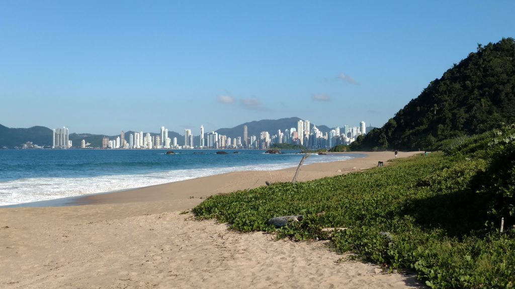 Praia com areia clara, mar azul e grama verde Conheça Balneário Camboriú