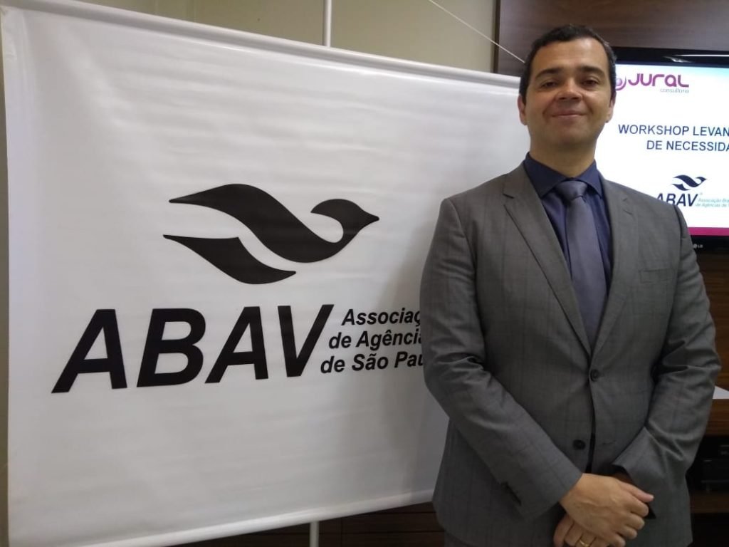 Homem de terno cinza ao lado de um banner branco Abav Expo 2019