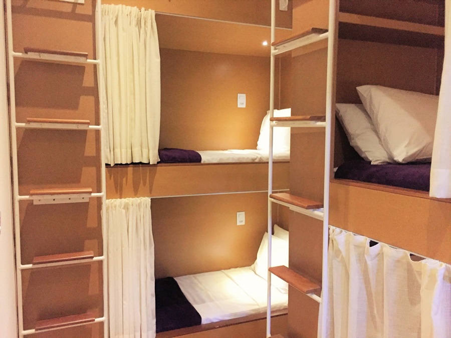 As cabines do Villa 25 Hostel com cortina, cama, cobertor roxo e lençol branco estendido Villa 25 Hostel