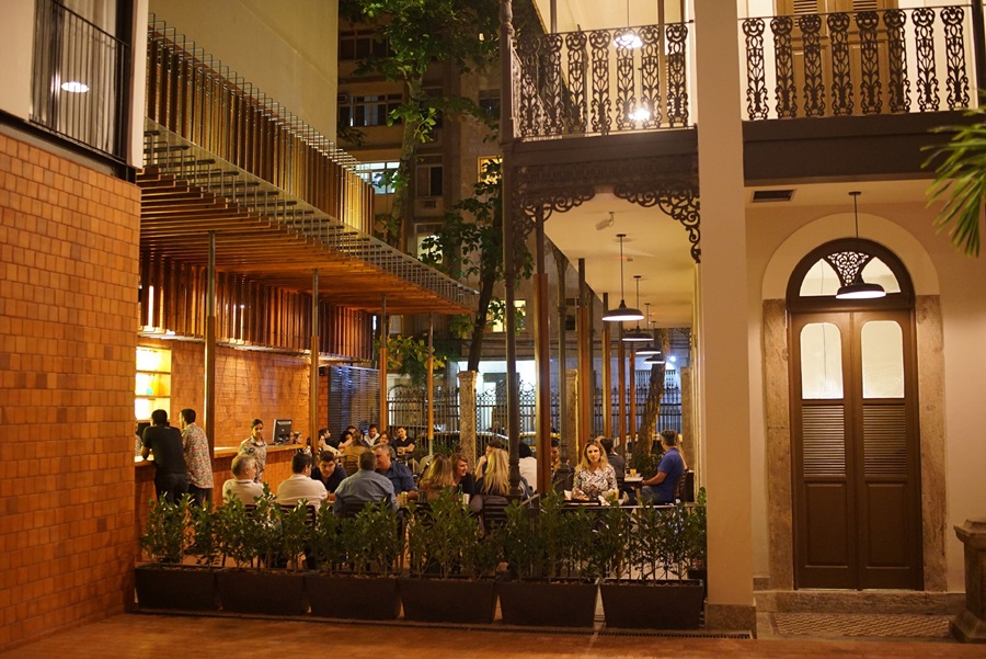 O ambiente do Villa Restobar com mesa, cadeiras, portas e pessoas sentadas e umas plantas a frente Villa 25 Hostel