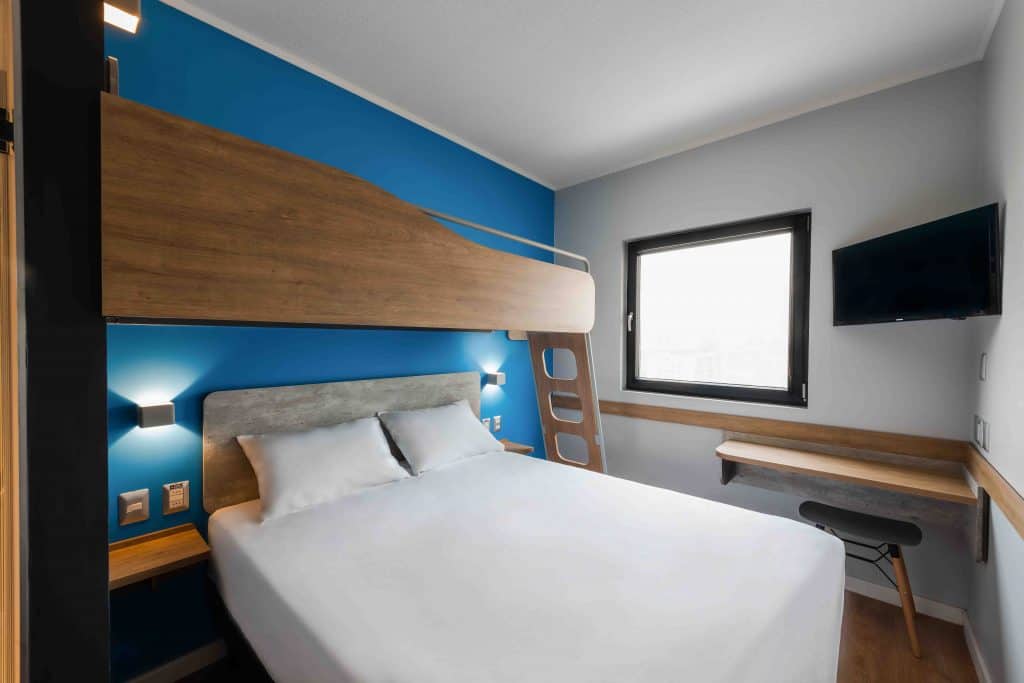 quarto de parede azul com móveis marrons, sendo uma cama de casal branca e outra cama em cima Ibis budget Providencia
