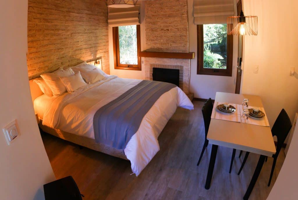 Um dos quartos do hotel com duas janelas, cama de casal, lareira, cadeiras e mesa O que fazer em Monte Verde