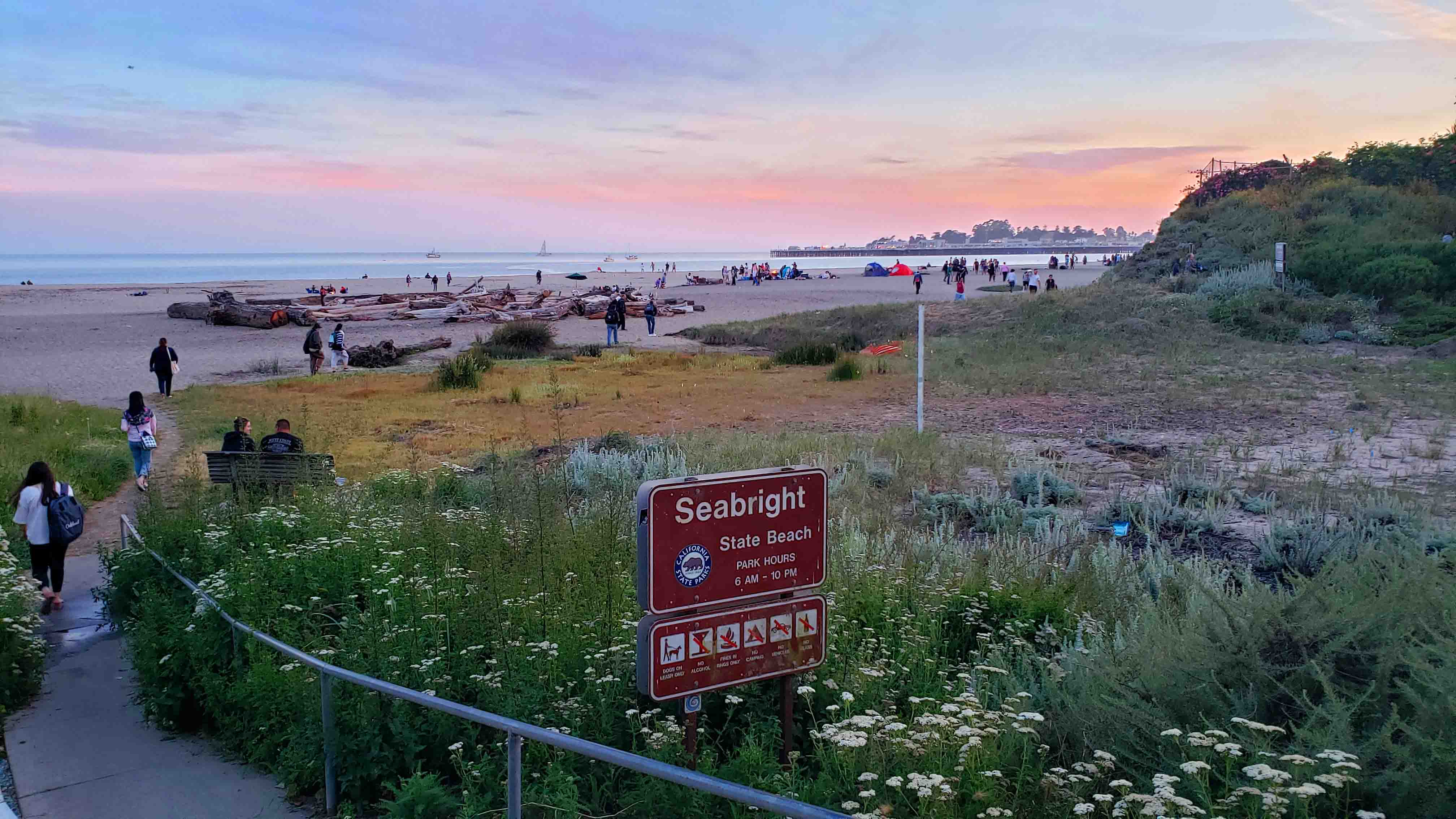 Caminho até chegar a praia com placa e muito verde. Ao fundo pessoas caminhando para praia Praias em Santa Cruz