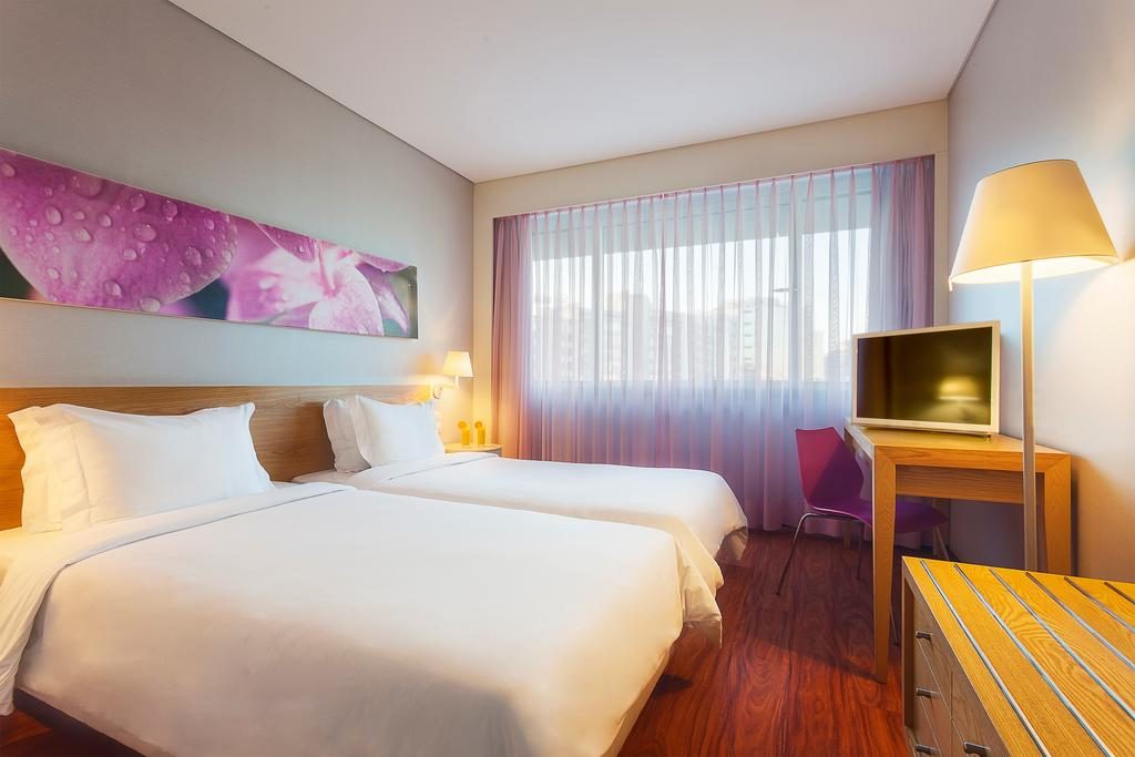 Um dos quartos do HF Fenix Garden com duas camas, mesa, cadeira, abajur, tv e decoração Onde ficar em Lisboa