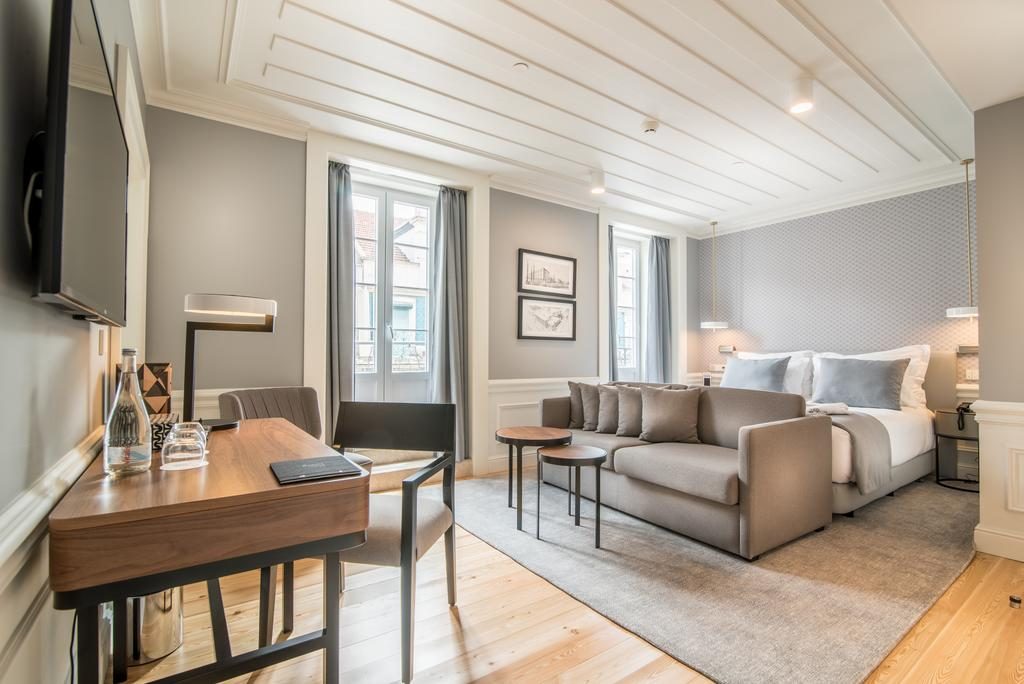 Um quarto de hotel com tv, cama, sofá, mesa, cadeiras, tapete e cortinas nas janelas Onde ficar em Lisboa
